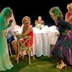 A bride in a veil and two women in dresses laugh together at a round table with a floral centerpiece. Another woman sits and smiles, while a man sits at the table in the background. The scene is set on grass with stage lighting.