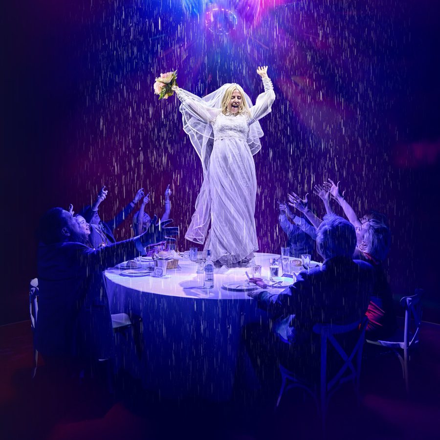 A person in a wedding dress stands on a table, arms raised, under theatrical rain while holding flowers. They are surrounded by people seated at the table, reaching up, amid dramatic purple and blue lighting.