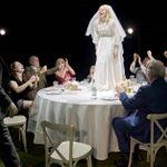 A bride wearing a white dress and veil stands on a round table, looking joyful. She is surrounded by seated guests who are clapping and cheering. The scene is festive, with plates and glasses on the table. The backdrop is dark, highlighting the bride.