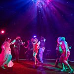 A group of people dancing energetically on a dimly lit stage under a disco ball. Vibrant pink, purple, and blue lights illuminate the scene, creating a lively atmosphere.