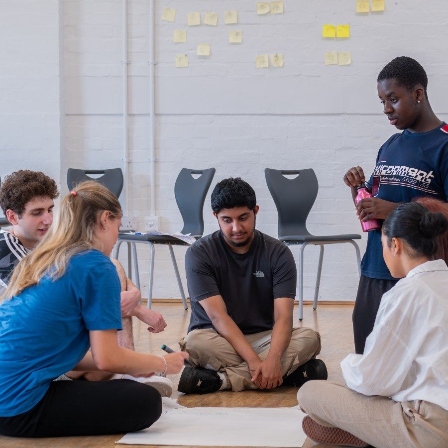 A group of five people sit in a circle on the floor, engaged in a discussion. One person is holding a pink water bottle. They appear to be collaborating on a project with papers in front of them. Chairs are lined up against a white wall in the background.