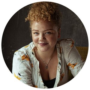 miranda-cromwell-headshot-1080x180 A woman with curly, light brown hair wearing a white blouse with orange floral patterns and a silver necklace smiles softly while seated indoors against a dark background.