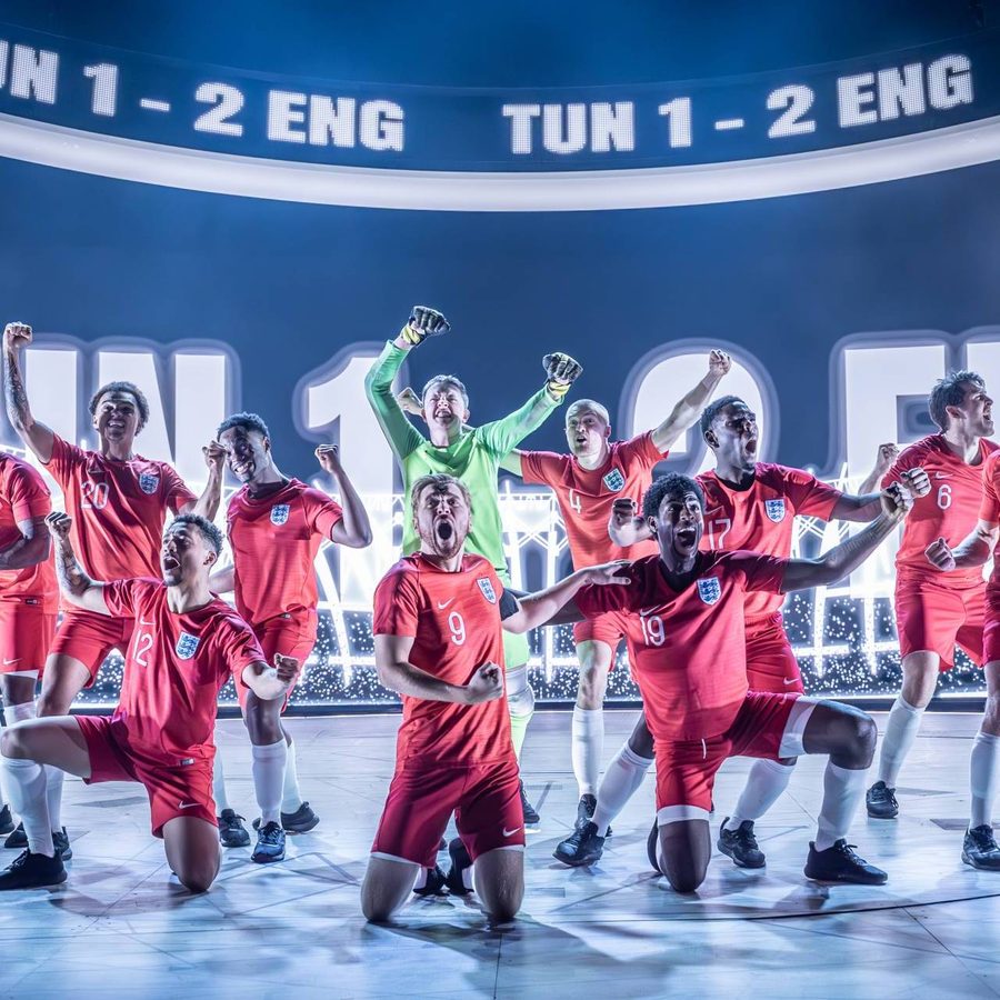 Football players in red football kits celebrate on stage under bright lights and a scoreboard showing 