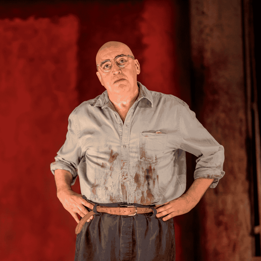 Two actors performing on stage in a dramatic scene. One actor, a bald man in a paint-stained shirt with his hands on his hips, stands confidently in the foreground. The other actor, a younger man in a white shirt, stands slightly behind him, looking thoughtful. Behind them is a large abstract red painting, evoking the style of Mark Rothko, with a dimly lit studio-like setting.