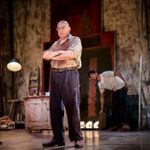 Alfred Molina stands center stage with arms crossed, his shirt stained with paint. In the background, a younger man bends to pick up an object near a large canvas featuring bold vertical blocks of red and black. The setting is a cluttered artist’s studio with distressed walls, art supplies, and industrial lighting.