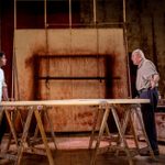 Alfrod Enoch and Alfred Molina stand on opposite sides of a large wooden table supported by sawhorses, engaged in an intense conversation. Behind them is a massive abstract canvas stained with deep red and brown hues. The setting is an artist’s studio, with worn tools, paint cans, and rustic furniture scattered around.