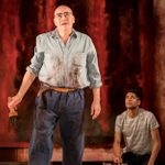 Alfred Molina stands in the foreground holding a paintbrush, while Alfred Enoch kneels beside him in the background. Behind them is a large abstract painting dominated by bold red hues.