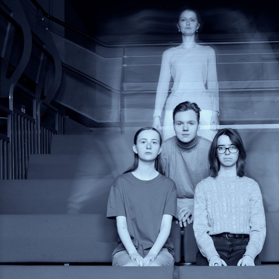Three serious young adults sit on theatre steps, while a ghostly, semi-transparent figure of a woman stands behind them, creating an eerie, surreal atmosphere.