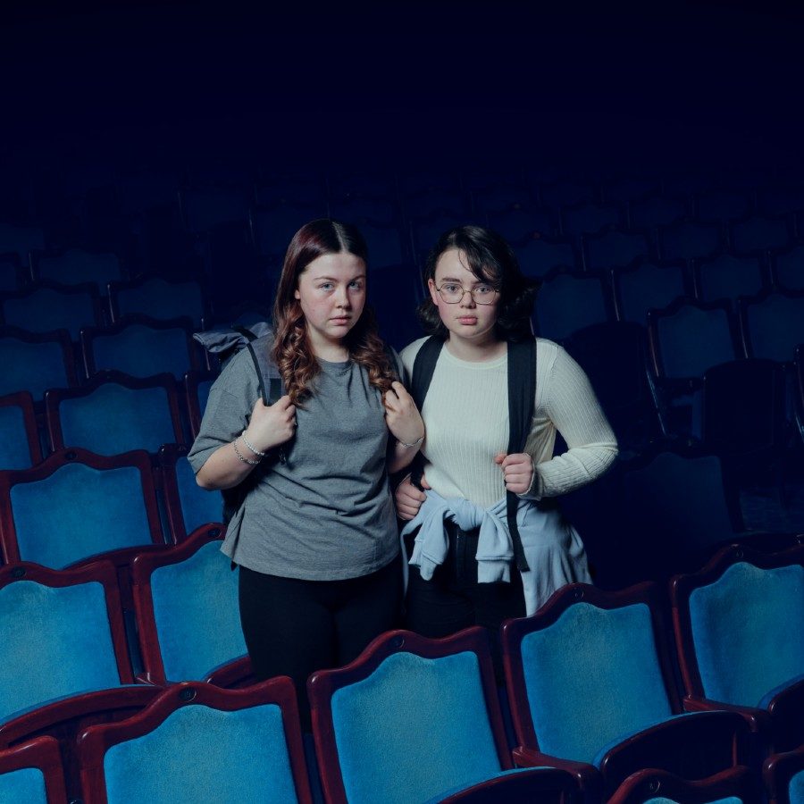 NTGDS_NF_Connections 2024_MiaAndTheFish_English_Digital_A4 Landscape_RGB_2000x1000 Two young people with backpacks stand in the middle of an empty theatre auditorium with blue seats. They have serious expressions.