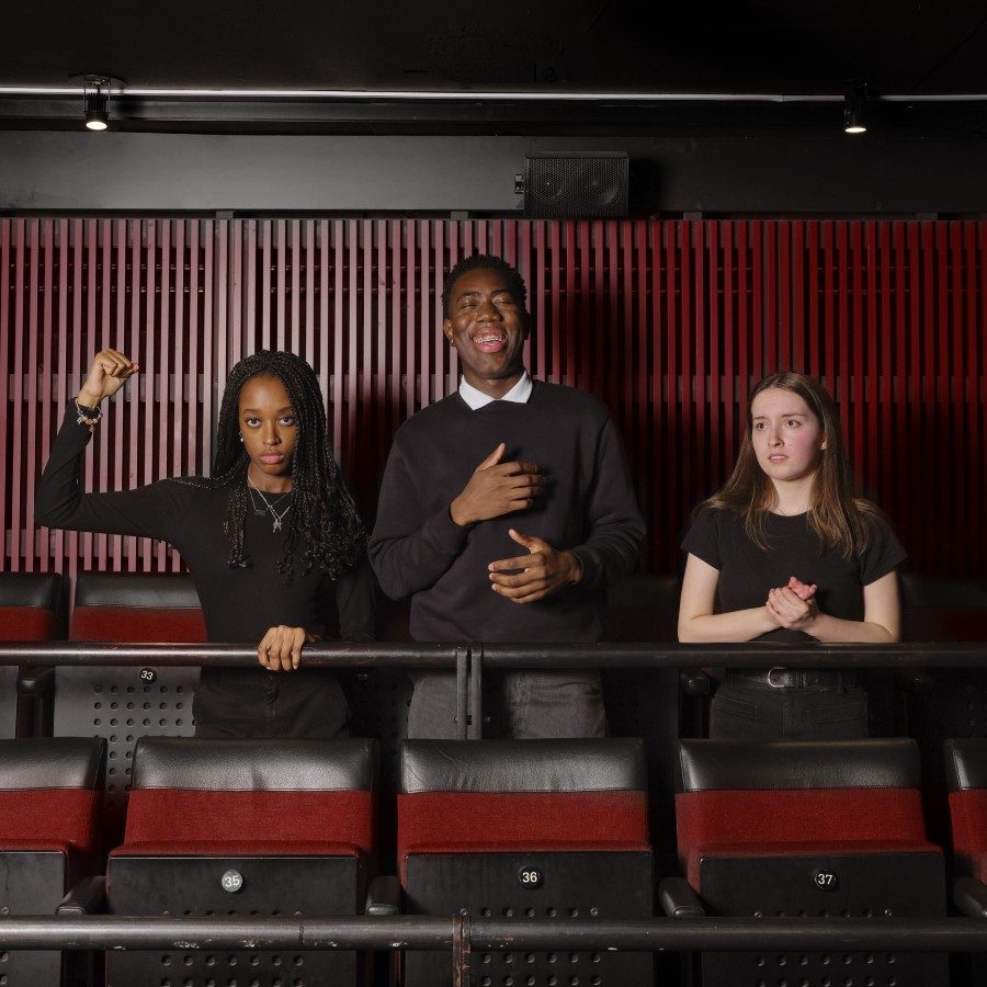 Three young people stand in a theatre against a dark wall. The person on the left raises a fist, the person in the centre has a hand on their chest and is smiling, and the person on the right clasps their hand. They are all dressed in black.