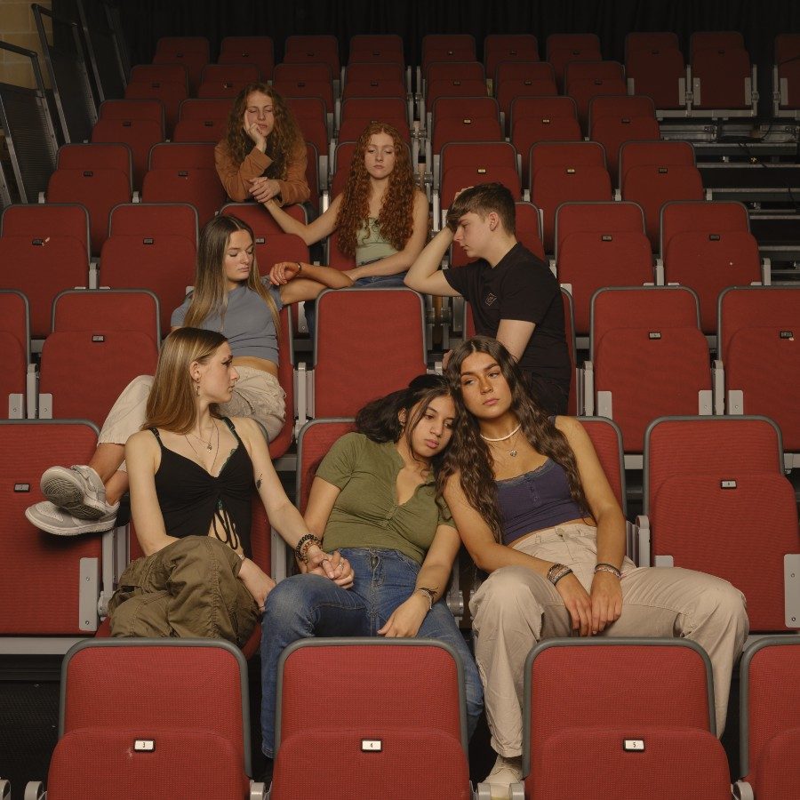 Seven young people sit scattered on red seats in a theatre auditorium. They look serious, some are holding hands.