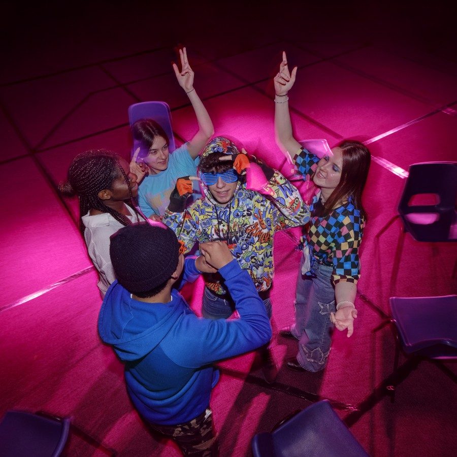 Five young people stand in a circle under pink and purple lighting. The central person wears blue plastic sunglasses. They are all wearing casual colourful clothing. A loose circle of plastic chairs surrounds them.