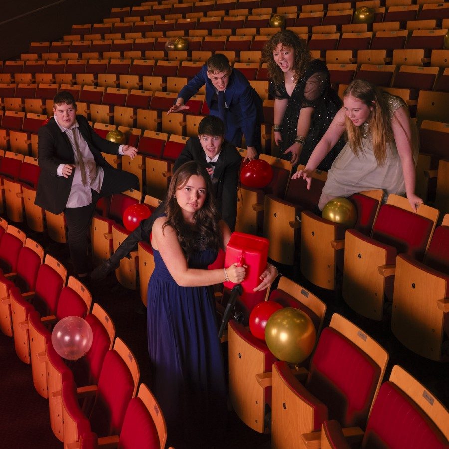 Six young people dressed in formal attire pose among rows of empty red theatre seats, scattered with balloons. One person in front holds a red plastic fuel can, while others reach toward it.