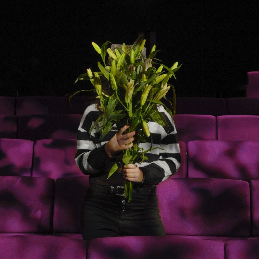 A person in dark trousers and a striped jumper stands between rows of empty purple chairs in a theatre. They are holding a large bouquet greenery that obscures their face.