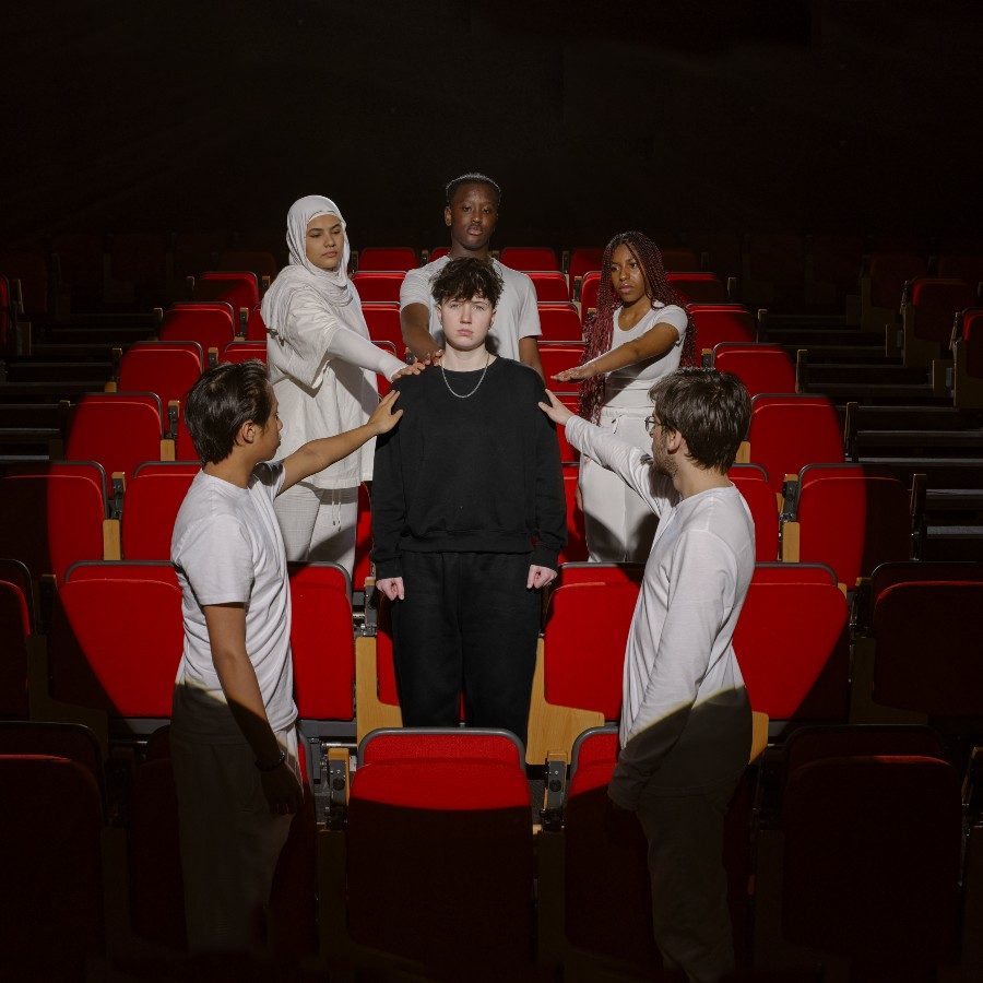 Six young people stand in a circle in a theatre auditorium with red seats. Five, dressed in white, encircle one person dressed in black, touching their shoulders. They are all lit in a spotlight with the rest of the auditorium in darkness.