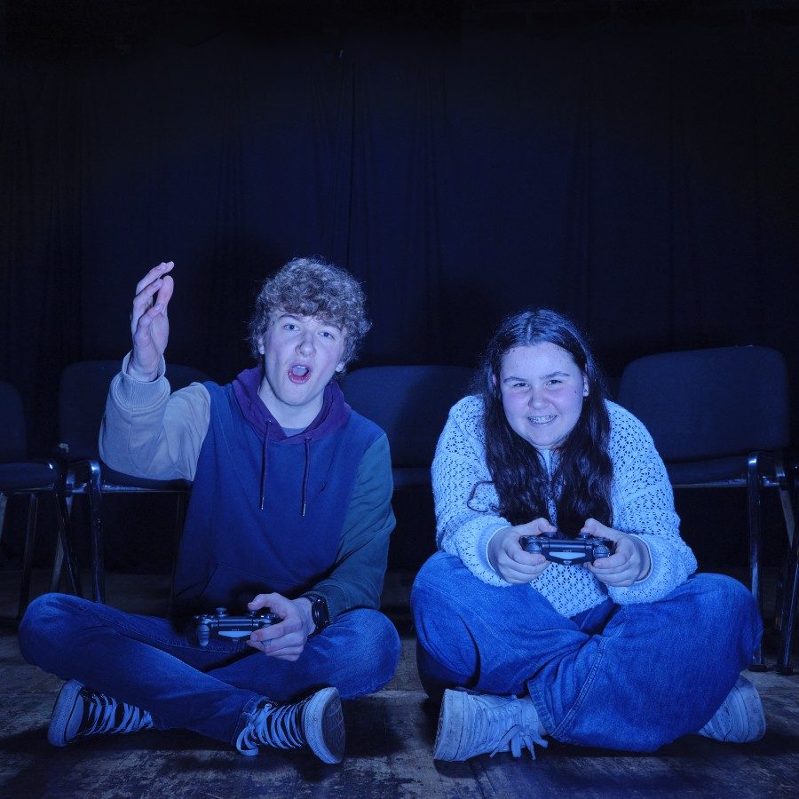 Two young people sit cross-legged on the floor in front of empty chairs, holding game controllers. The person on the left gestures animatedly, while the person on the right smiles. The scene is lit with blue light.