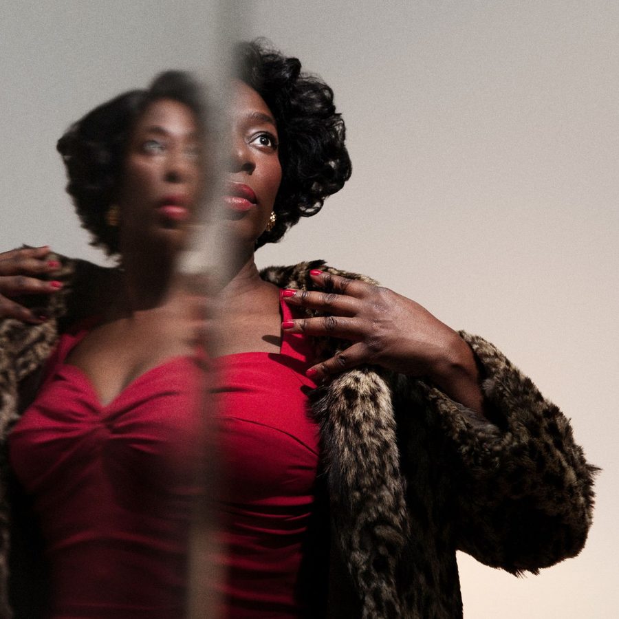 Trouble-in-Mind-National-Theatre-2021-masthead A woman with curly hair and red lipstick gazes upward, her reflection blurred beside her. She wears a red top, gold earrings, and a fur-like coat, with hands touching her collar. The background is neutral.