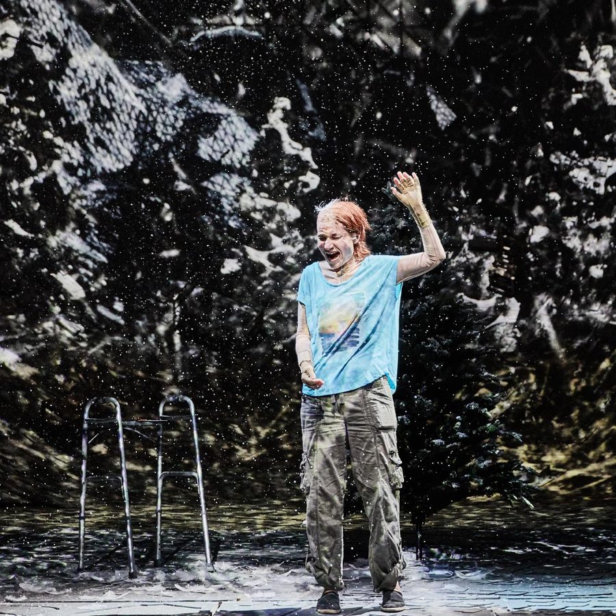 A person stands on a dark stage, wearing a blue t-shirt and cargo pants, with their hand raised. A walker is beside them, and video projections cover everything on the stage.