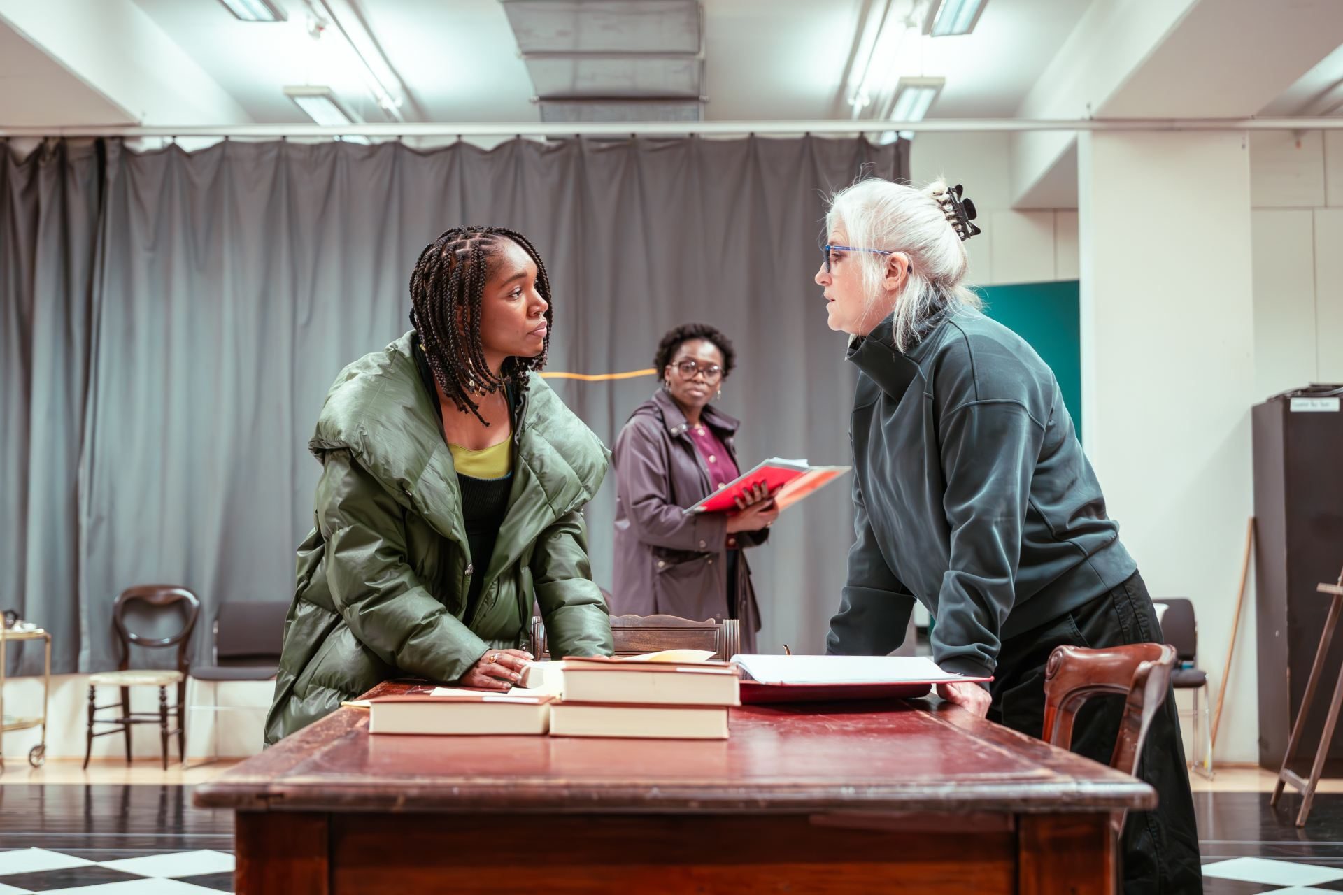 Three actresses rehearsing a scene. Two are leaning over a table with books, talking seriously, while the third stands in the background holding a red notebook and watching them.