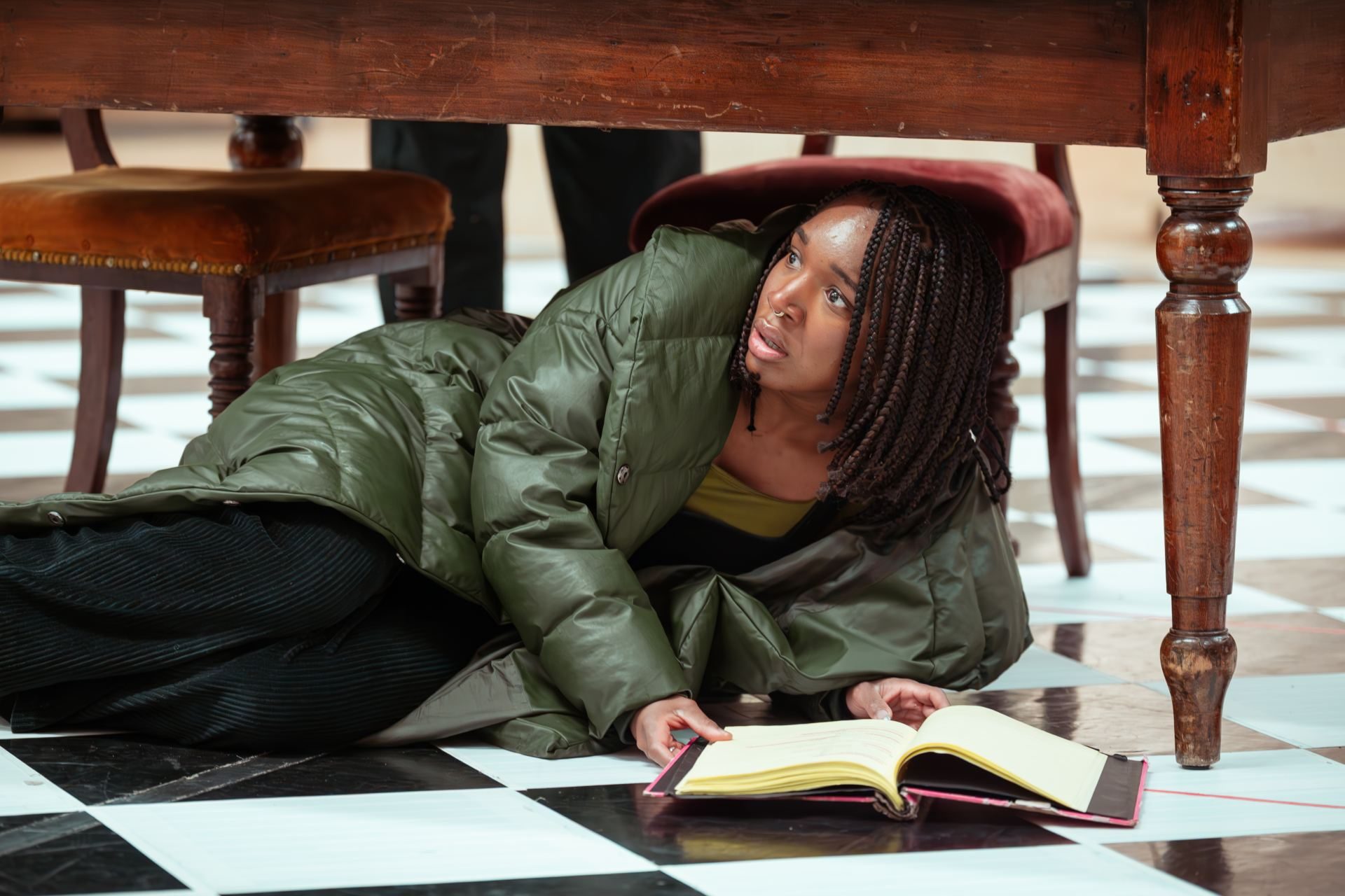 An actress in a green coat lies on a black-and-white tiled floor under a table, looking up with a concerned expression. An open book is on the floor in front of her.